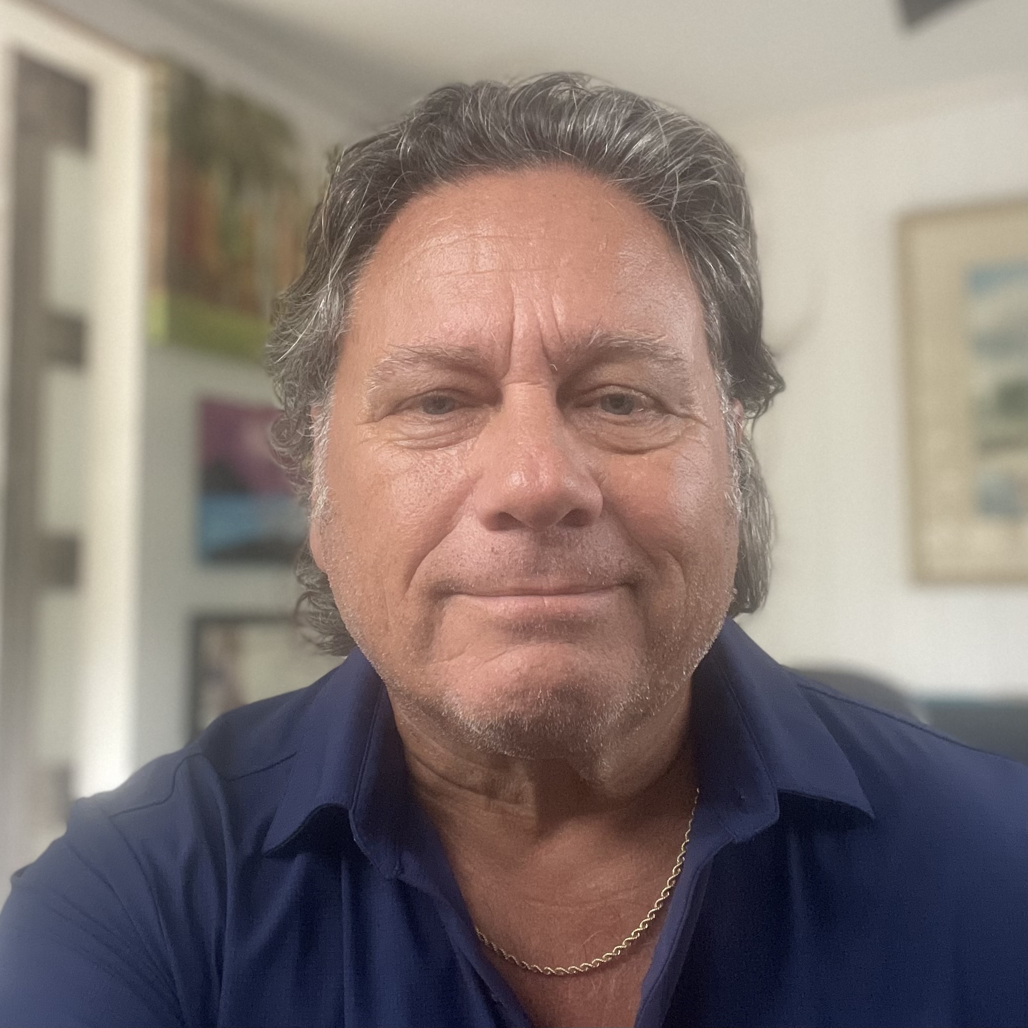 Smiling man with gray hair in a blue shirt, wearing a gold chain. He's indoors with blurred wall art and furniture in the background, creating a cozy atmosphere.